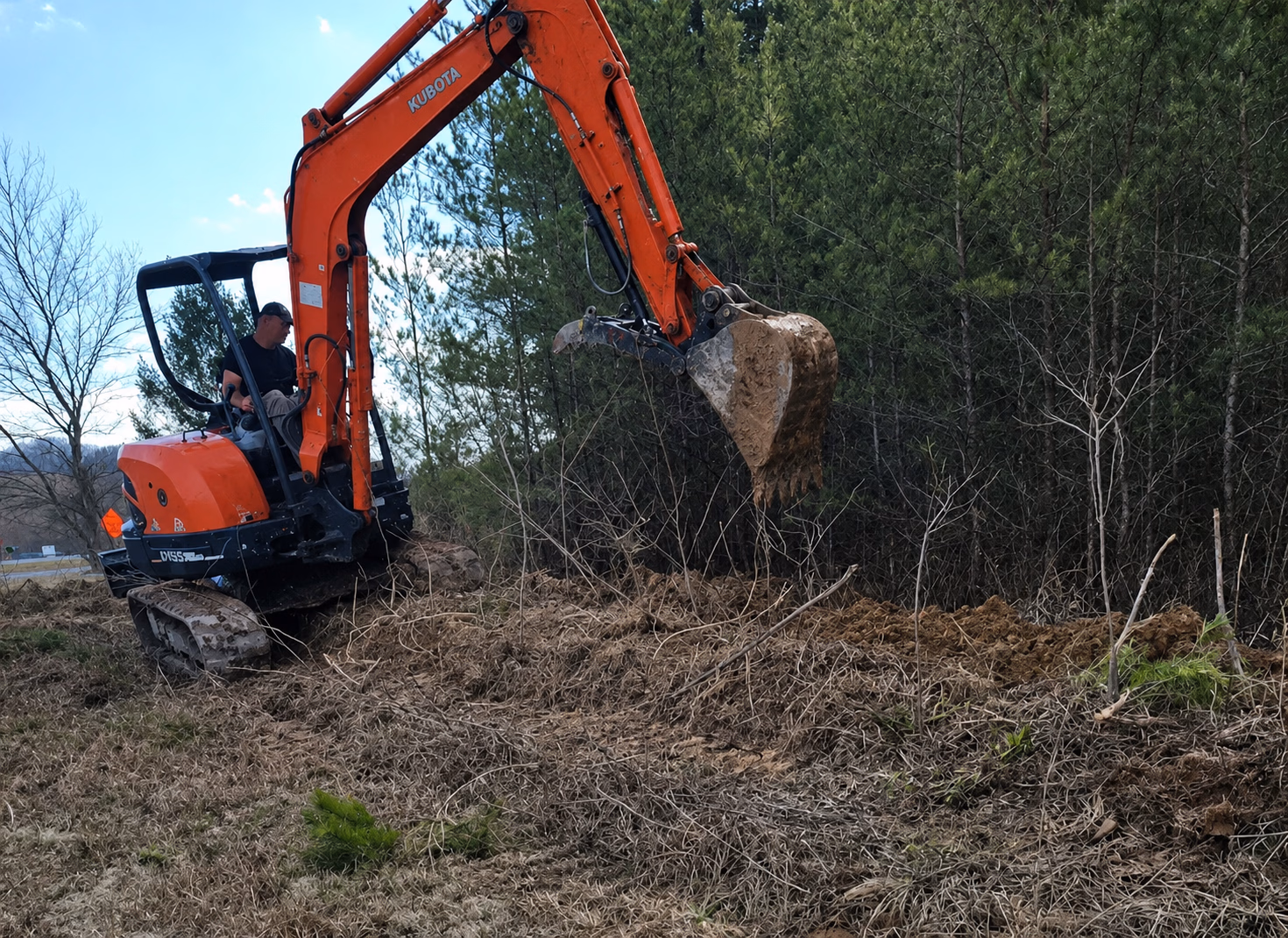 Man operating orange Kubota excavator digging soil near a forest with pine trees.