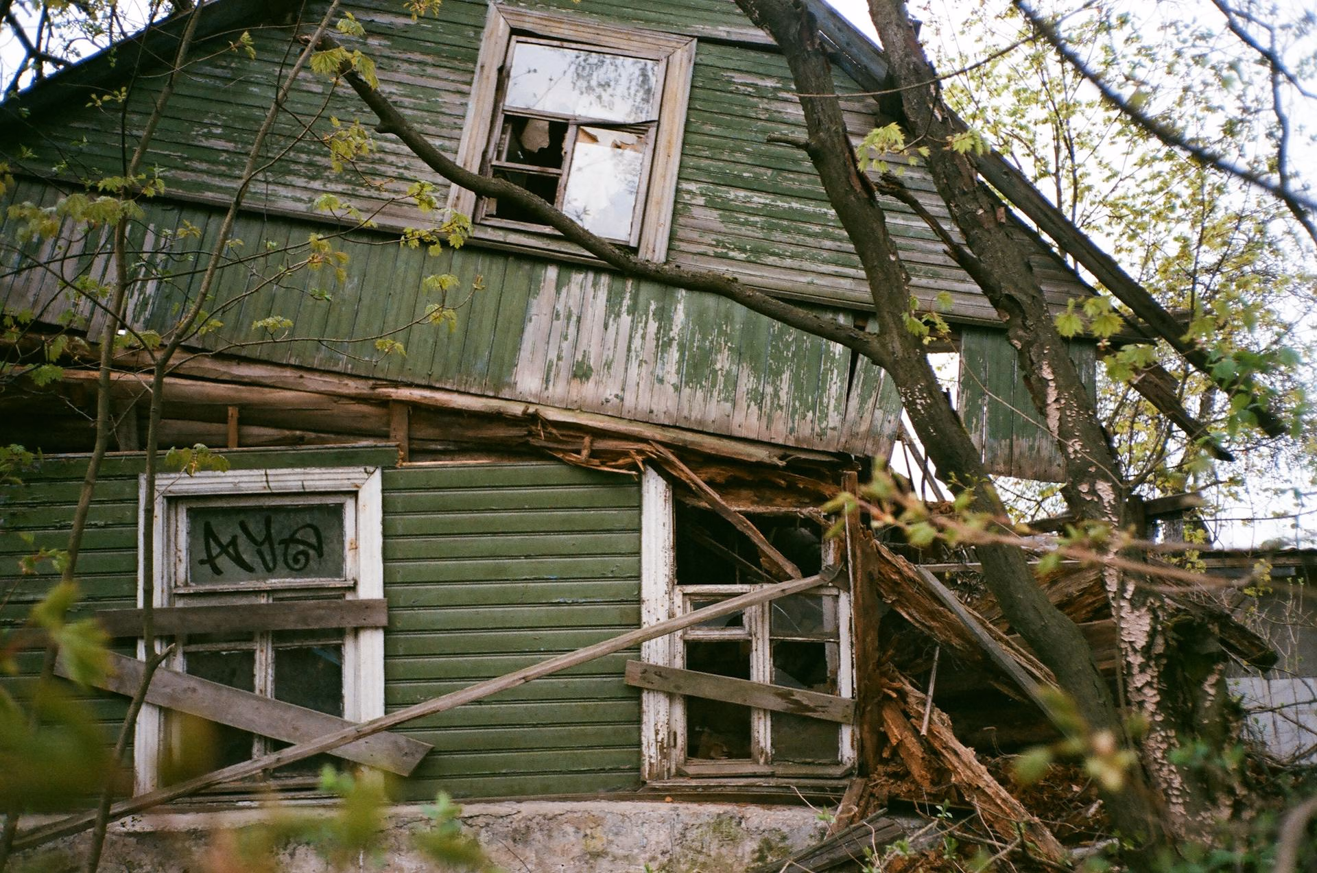 Old green wooden house with broken windows and boarded-up sections, surrounded by trees.