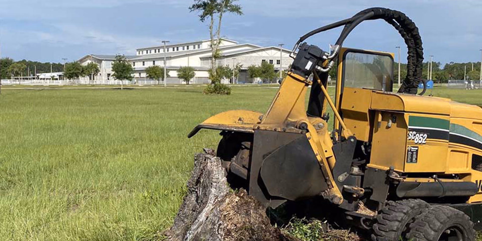 Yellow stump grinder machine cutting a tree stump in a grassy field near a large building.
