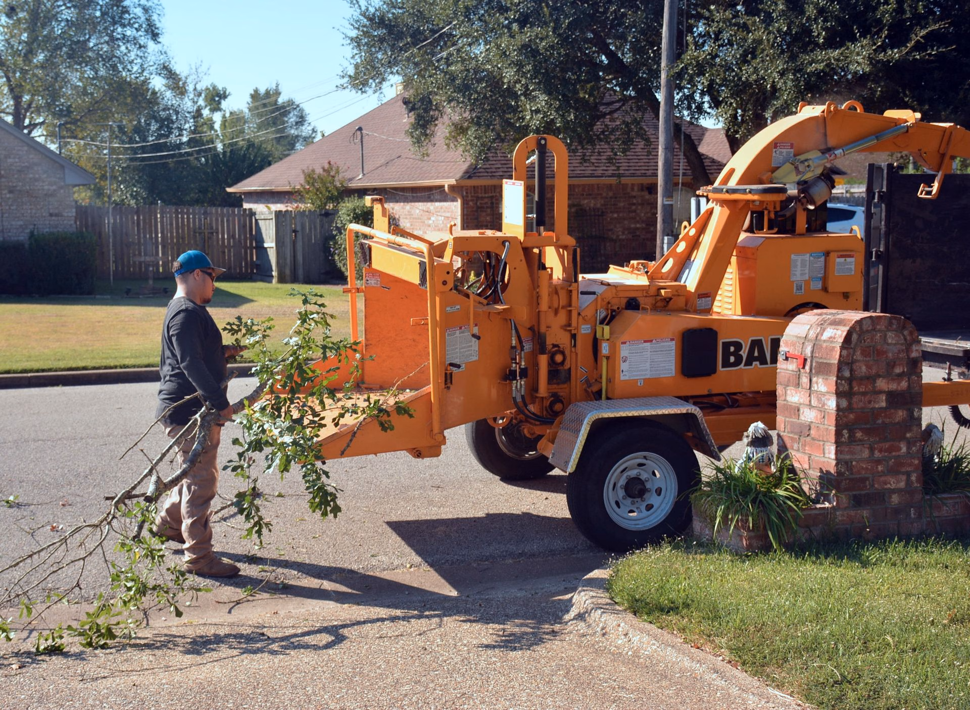 Worker feeding tree branches into an orange wood chipper in a suburban driveway.