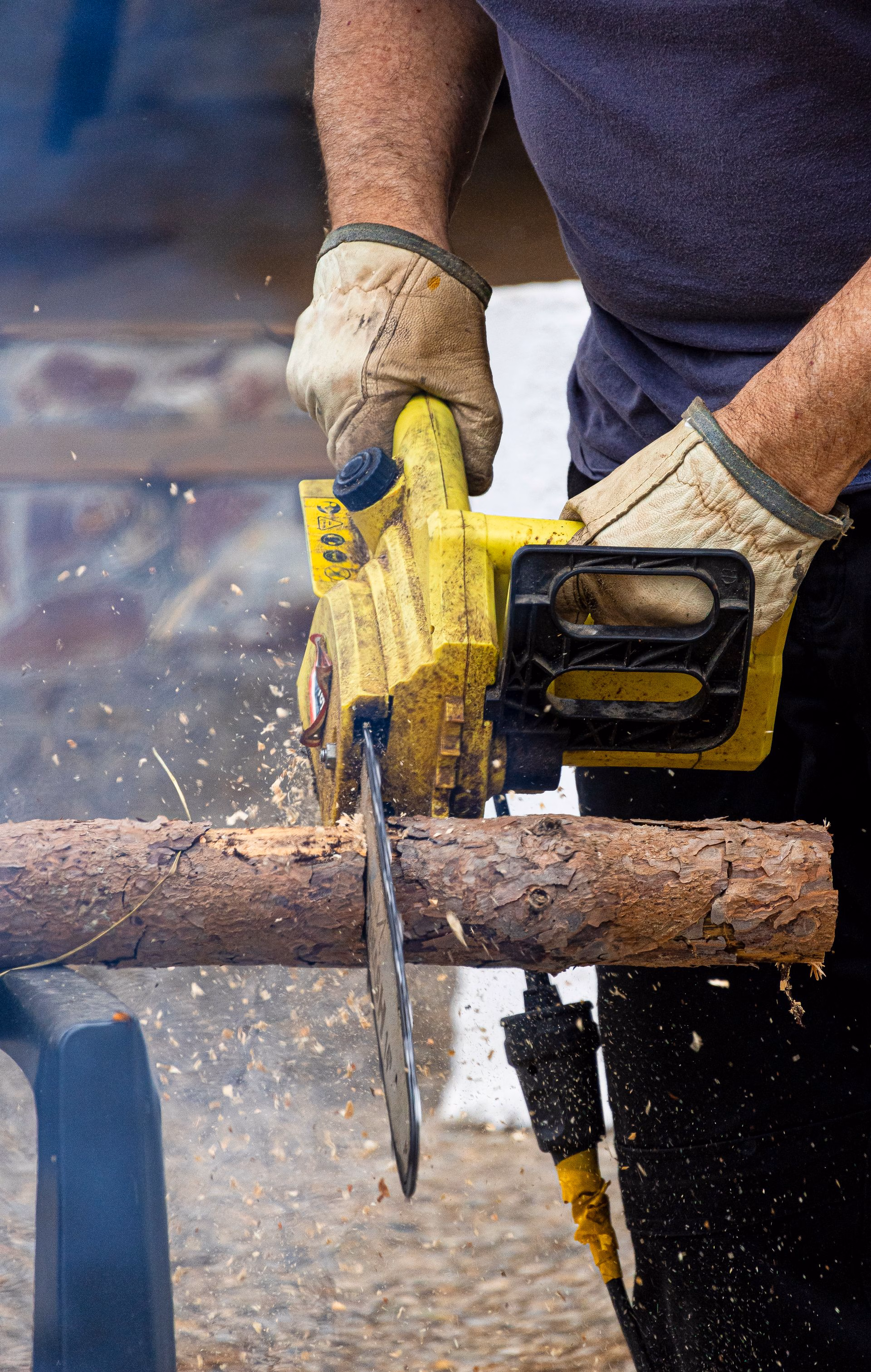 Person wearing gloves using a yellow chainsaw to cut a log, sending wood chips flying.